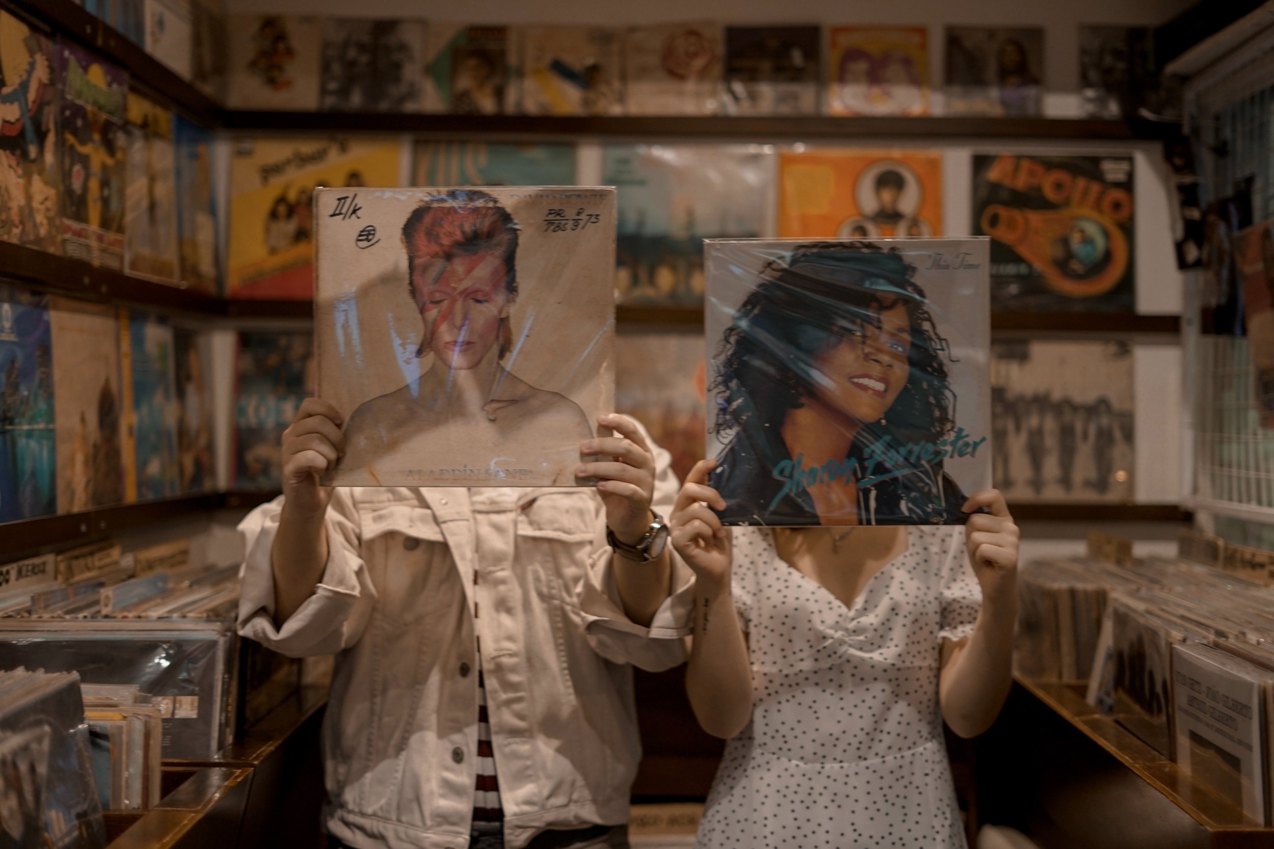 Two people stand in a record store holding up record albums that cover their face.