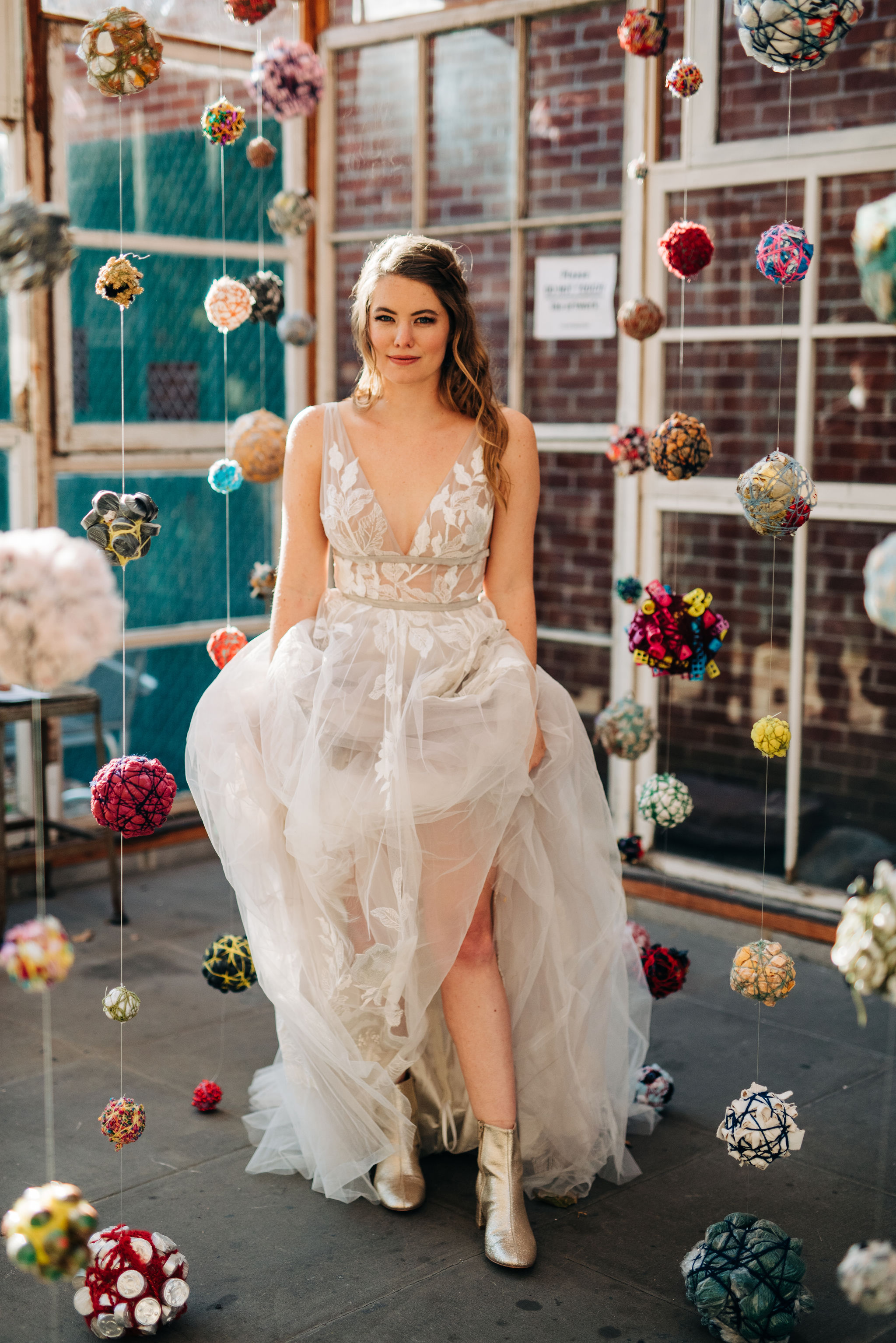 A photo of a white woman with brown hair in a wedding dress standing in a room full of pom poms hanging from the ceiling.