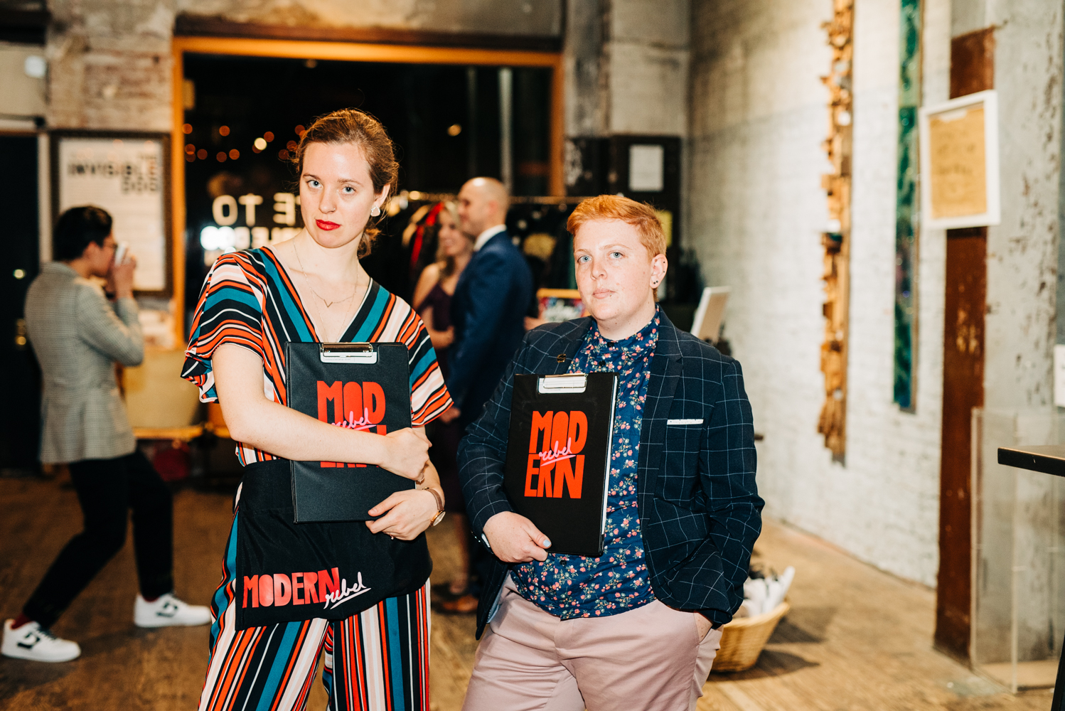 Two white people in fancy outfits stand in a room holding clipboards that say Modern Rebel on them, they are looking at the camera with serious faces.