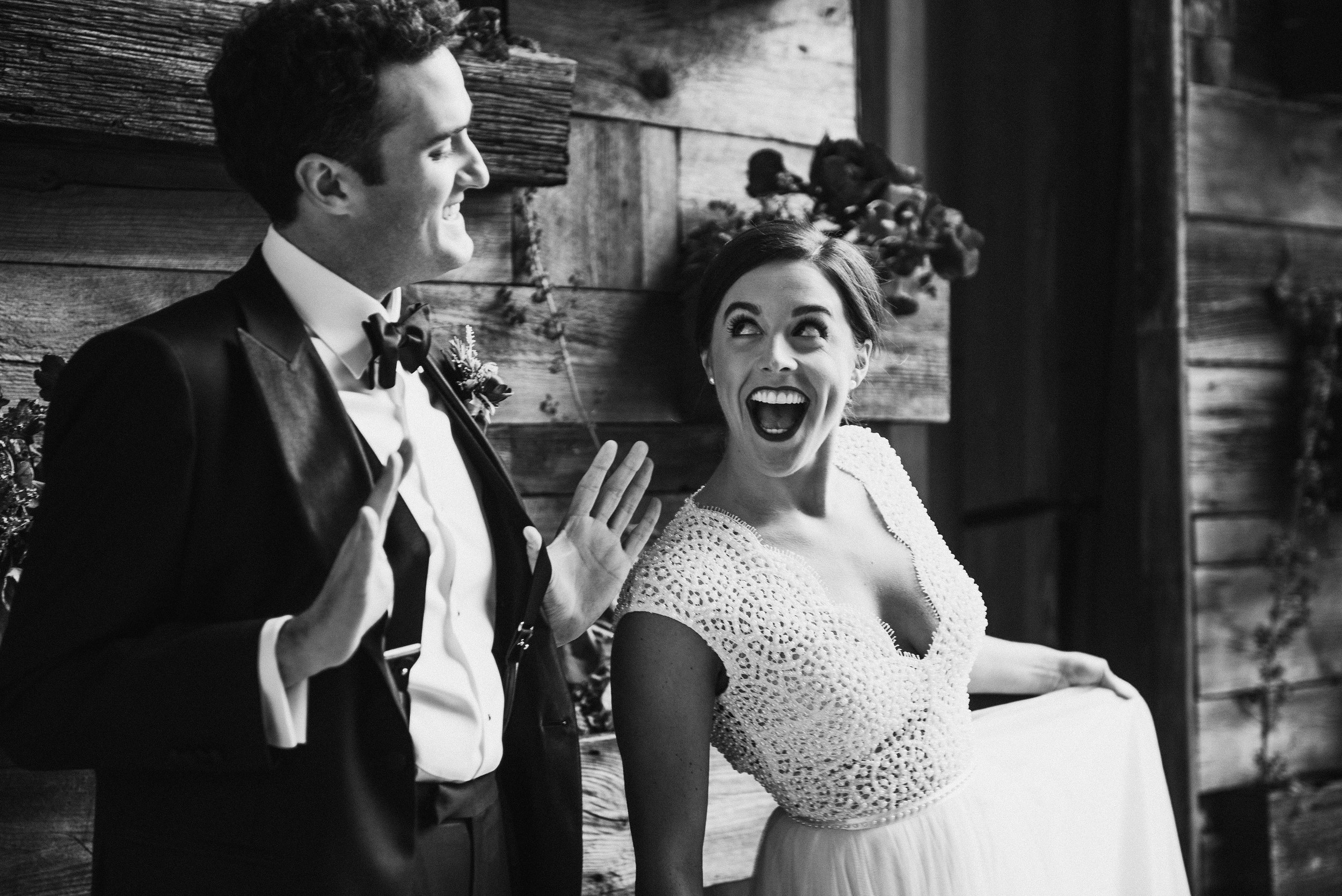 Two marriers stand against a wooden wall, one in a suit holding their suspenders out, the other in a wedding dress smiling big.