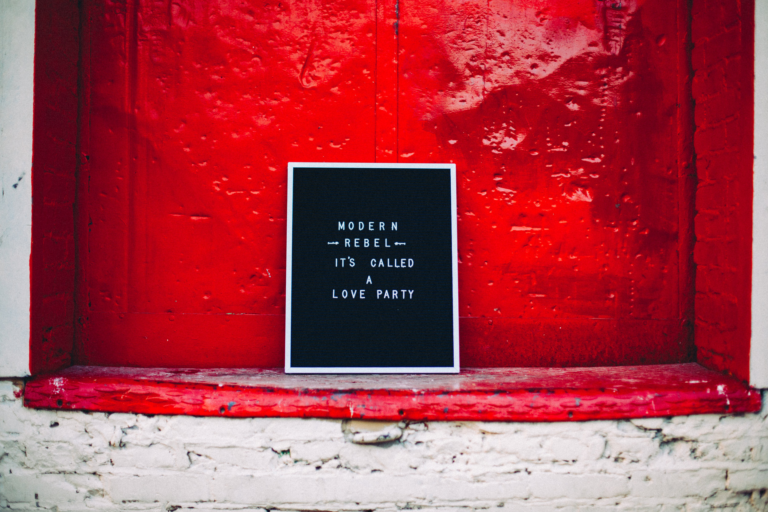 A letterboard sign saying "Modern Rebel - it's called a Love Party" sitting against a bright red doorway.