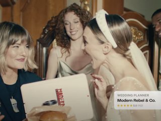 A wedding planner hands a hamburger to a marrier in a wedding dress