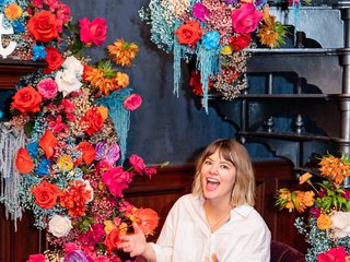A white woman with brown hair sits in a chair in front of a wall of flowers, smiling wide with her hands outstretched