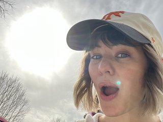Amy, a white woman, wears a hat and looks at the camera while the sun shines in the sky above her