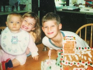 Three young kids gather at the table around a gingerbread house