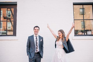 Two people in wedding attire stand in front of a wall, one person has their arms outstretched in peace signs