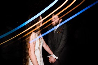 Two people hold hands on their wedding day in a dark room with streaks of colored light over their faces