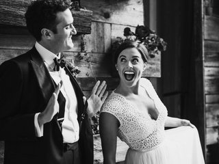 A man and a woman in wedding attire lean against a wall smiling at each other