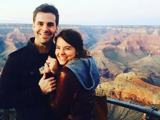 Two people stand holding hands and smiling in front of the grand canyon