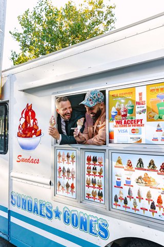 Two white men lean out of an ice cream truck holding ice cream cones smiling