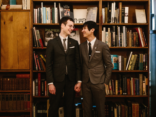 Two grooms stand in front of books in a bookstore smiling at each other