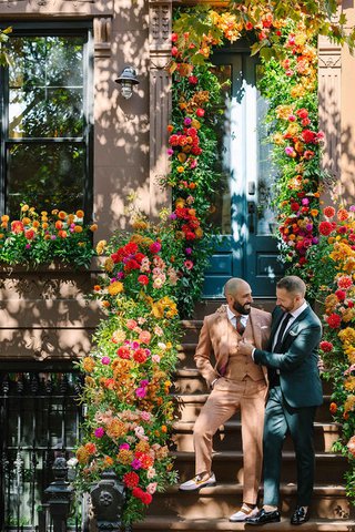two white men stand lovingly together in front of a brooklyn brownstone building that is decorated with flowers