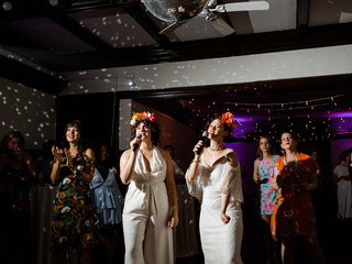 Two queer women brides in white attire sing karaoke under a disco ball