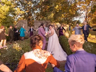 A group of people at a wedding stand in a circle outside on grass holding hands surrounding two marriers