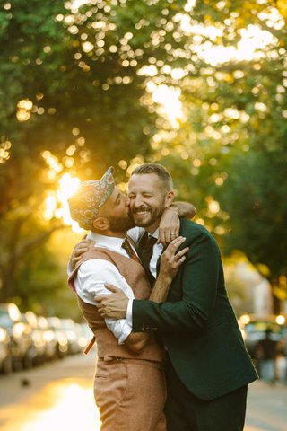 Two white men stand in the street with the sun behind them, one is kissing the other's cheek