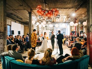 Two people get married by two officiants in a room surrounded by loved ones.
