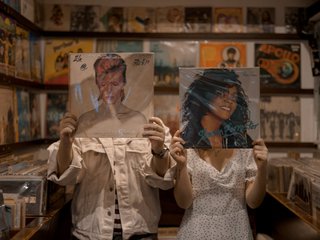Two people stand in a record shop covering their faces with record albums