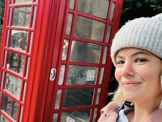 A white woman in a hat and jacket smiles in front of a red British phone booth