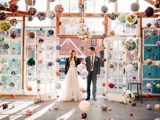 two people in wedding attire stand in a room full of colorful pompoms hanging from the ceiling